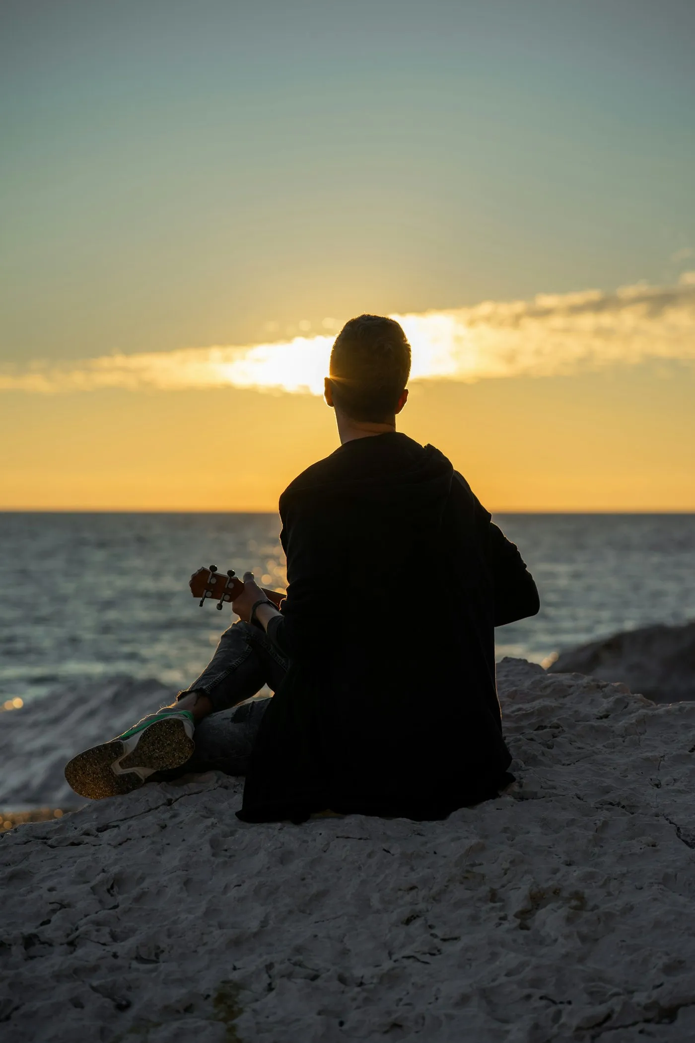 Un músico de espaldas, sentado en la arena con su guitarra, mirando el mar al atardecer